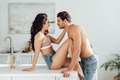 Couple smiling, looking at each other and sexy girl sitting on table touching man in kitchen - PhotoDune Item for Sale