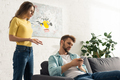 Angry woman standing near boyfriend with smartphone on couch - PhotoDune Item for Sale