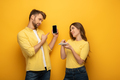 Young couple pointing on smartphones while looking at each other on yellow background - PhotoDune Item for Sale