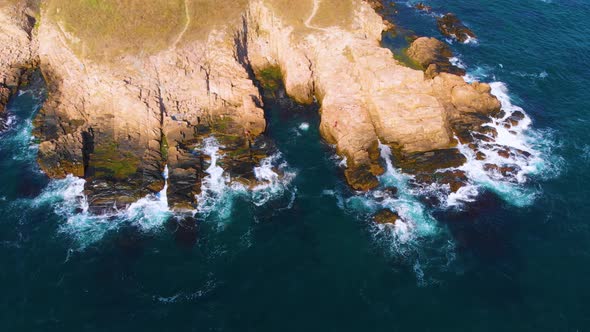 Aerial footage of a person jumping off a cliff in the sea and then swimming. Waves are crashing into