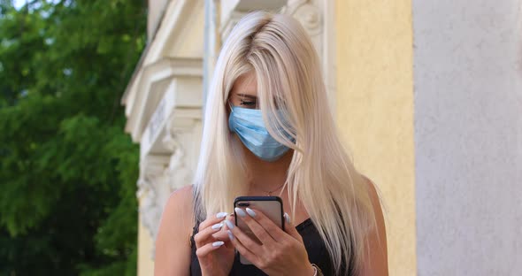 Young Blonde Woman in Protective Medical Mask Walks Down To the Street Uses Phone Texts Scrolls