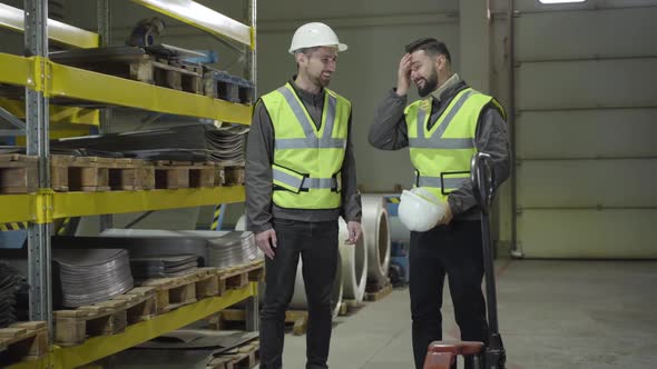 Two Smiling Caucasian Men Standing at Steel Metal Warehouse and Talking. Workers Doing Their Job at