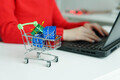 Shopping cart with gifts on table. Woman using laptop on the background. Shopping online in holidays - PhotoDune Item for Sale