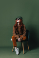 Stylish redhead woman in sunglasses and ginger shirt and pants sits in chair on green background - PhotoDune Item for Sale