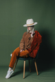 Stylish blond hair woman in hat nad orange pants sits in chair on green backgorund in studio - PhotoDune Item for Sale
