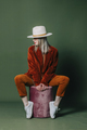 Stylish blond hair woman in hat and orange pants sits ottoman on green backgorund in studio - PhotoDune Item for Sale