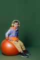 Stylish little boy in headphones sits on orange swiss ball on green background - PhotoDune Item for Sale