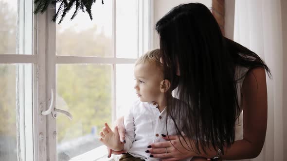 Young Mother is Sitting with Her Son on the Window Sill Decorated with Christmas Wreath and Looking