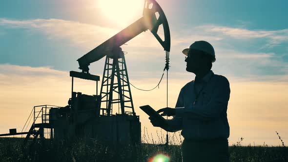 Dark Silhouette of Engineer with Tablet with Oil Pumpjack Working in Background