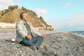 Bearded man sitting in lotus pose on pebble beach,meditating and listening music in headphones - PhotoDune Item for Sale