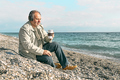 Bearded man listening music in headphones and drinking coffee while sitting on the beach. - PhotoDune Item for Sale