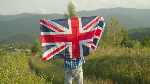 Joyful Man Standing with Big Flag of Great Britain on Top of Mountain at Sunset