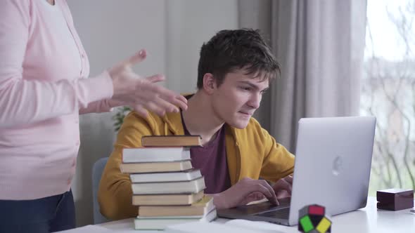 Young Caucasian Brunette Boy Playing on Laptop As His Mother Coming Up with Stack of Books. Woman