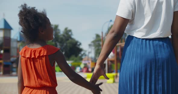 Back View of African Little Girl Hold Hands with Mother in City Park