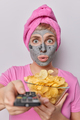 Surprised woman stares impressed at camera holds remote control watches TV at home eats crisps - PhotoDune Item for Sale