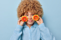 Horizontal shot of cute curly haired young woman holds small orange flowers near cheeks looks - PhotoDune Item for Sale