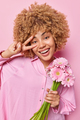 Positive carefree woman with curly hair makes peace sign over eye smiles broadly holds bouquet of - PhotoDune Item for Sale