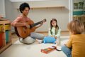 Happy black kindergarten teacher playing acoustic guitar to group of kids. - PhotoDune Item for Sale