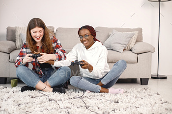 a joystick. Black girl wearing eyeglasses and beige sweater, caucasian girl wearing plaid shirt. Girls playing games together.