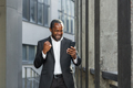 Happy young african american man near office. He holds the phone in his hands, celebrates, rejoices - PhotoDune Item for Sale