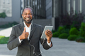 A young African American man in a suit is standing outside wearing headphones, holding a tablet - PhotoDune Item for Sale