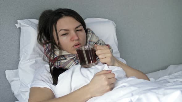 Sick Woman Lies in Bed Wrapped in a Scarf and Drinking Herbal Tea