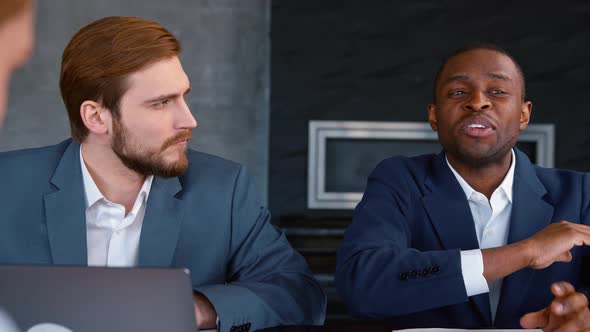 Young african american man in suit telling idea to colleagues while sitting at the desk in office