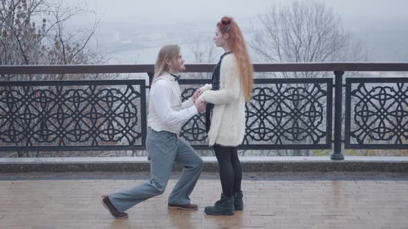 Side View of Stylish Boy in Elegant Retro Style Clothes Bending Down on One Knee and Proposing To