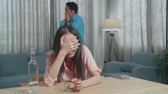 Depressed Woman Pouring Vodka In A Shot Glass Before Drinking And Crying, Man Walking Behind Her