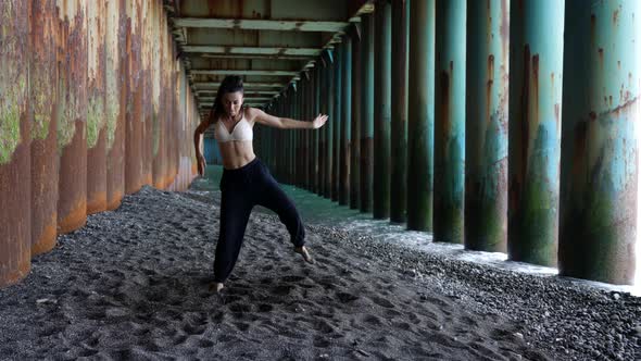 a Barefoot Woman Dances Under the Pillars of the Bridge Against the Background of the Incoming Waves