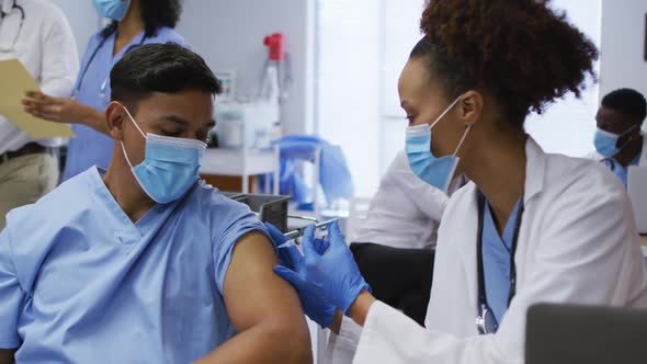 Mixed race female doctor giving covid vaccination to male colleague in hospital, both in face masks