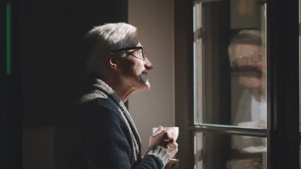 Senior Man Standing and Looking Out of Window Drinking Coffee
