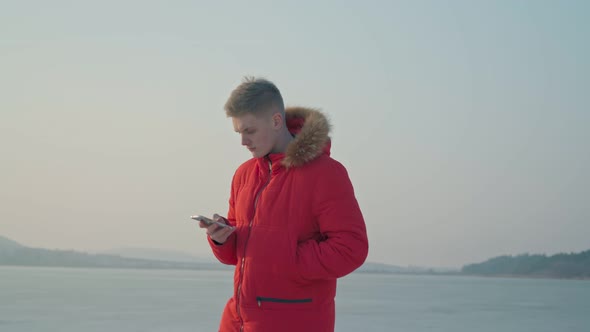 A Young European Guy Walks Along the Lake with a Smartphone Communicating on Social Networks