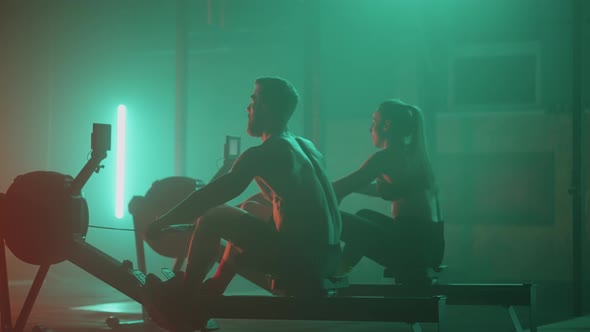 A Man and a Woman Perform Strong Athletes Perform Exercises in the Gym on a Rowing Machine in Slow