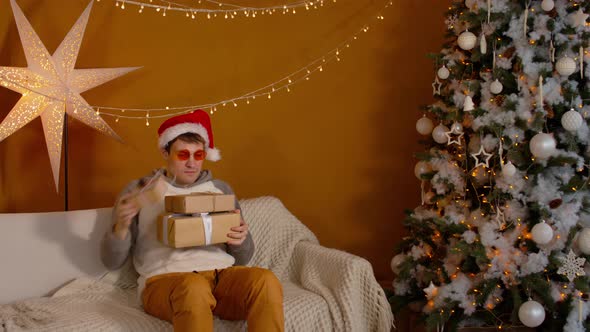 Man in Santa Hat and Bright Glasses with Gift Boxes Sitting on Couch Near Christmas Tree