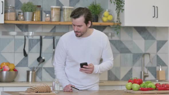 Young Man Making Online Payment on Smartphone in Kitchen