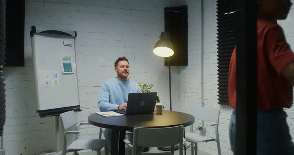 A Young Woman Enters the Meeting Room Where Her Colleague is Working on a Laptop