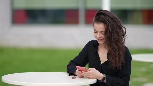 Adult Beautiful Brunette Woman is Sitting at Table of Street Cafe and Using Smartphone