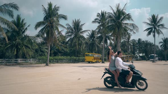 Girl Guy Ride Scooter on Square Against Tropical Trees