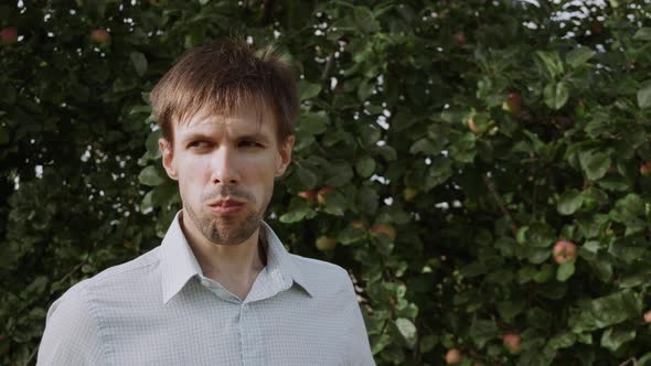 Man Nibbles Piece Apple and Chews It Near Green Apple Tree Closeup