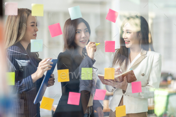 Young creative business Asian woman standing with her colleagues writing new ideas on sticky notes o