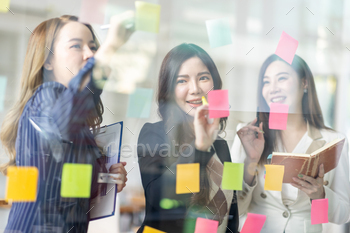 Young creative business Asian woman standing with her colleagues writing new ideas on sticky notes o