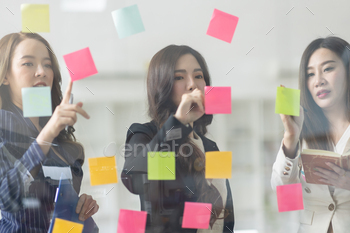 Young creative business Asian woman standing with her colleagues writing new ideas on sticky notes