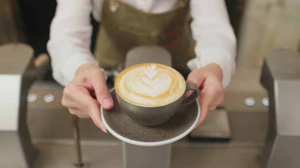 Close up of Asian barista woman holding latte coffee artwork hot cup giving to customer in cafe.