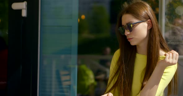 Cheerful Young Woman with Trendy Sunglasses Is Drinking Coffee in Street Cafe, Looking on Phone