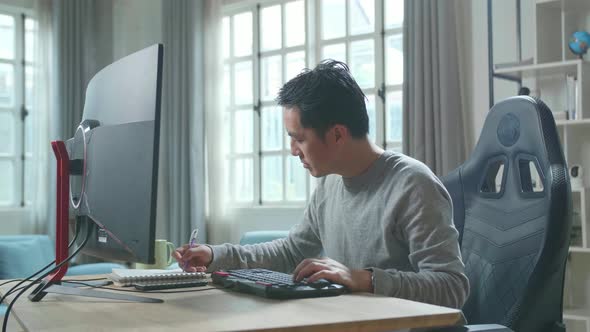 Asian Man Looking At Desktop Computer And Writing In Notebook While Working At Home