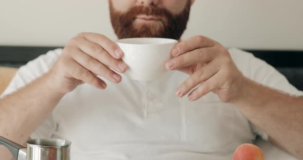 Happy Young Man Smelling His Coffee in Bed and Smiling. Cheerful Bearded Guy Having Breakfast While