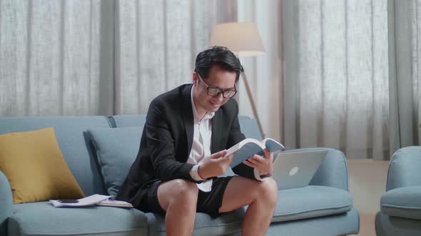 Asian Businessman In Jacket And Shorts Reading A Book After Working With A Laptop At Home