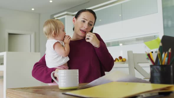 Caucasian mother holding her baby and talking on smartphone while working from home