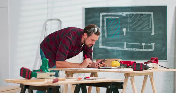 A Handsome Carpenter in a Flannel Shirt Counts the Dimensions on a Smartphone in a Carpentry